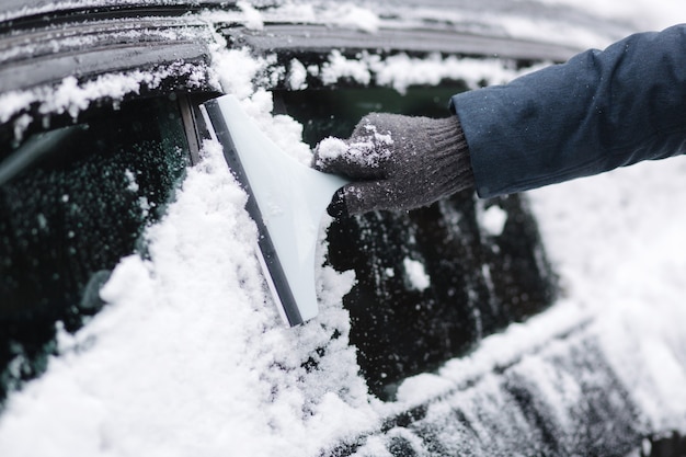 男のクローズアップは、スノースクレーパーで車の雪に覆われた窓を掃除しています。スクレーパーに焦点を当てます。コールド