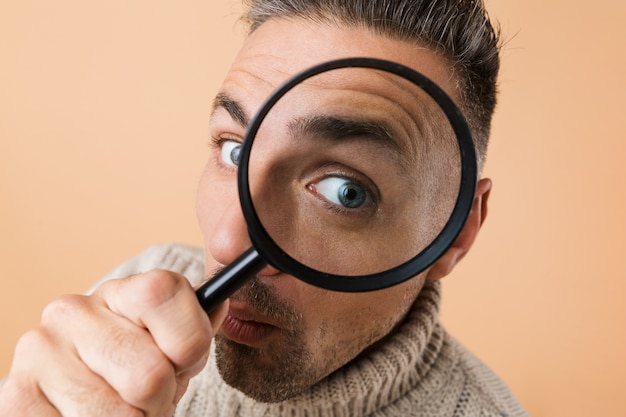 Close up of a man looking through a magnifying glass isolated over beige wall