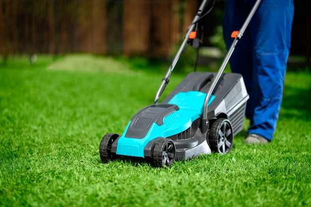 Close-up of a man in overalls with a lawn mower cutting green grass in a modern garden. 