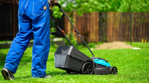 Close-up of a man in overalls with a lawn mower cutting green grass in a modern garden. 