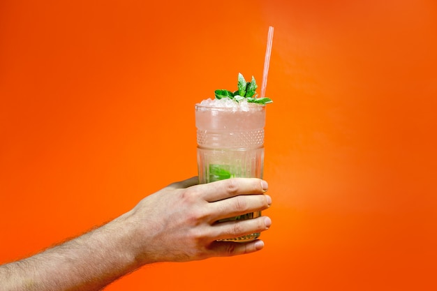 Close up of a man's hand holding a glass of tropical mojito cocktail on orange surface with copy space. Summer time vacation concept.