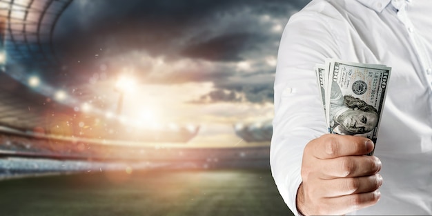 Close-up of a man's hand holding US dollars against the background of the stadium. The concept of sports betting, making a profit from betting, gambling. American football.