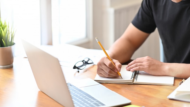 Hs dell'uomo del primo piano che scrive sul computer portatile della carta del taccuino.