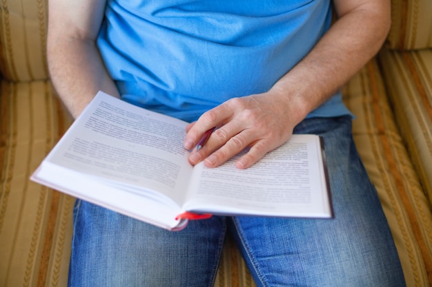 Chiuda su dell'uomo che si siede sullo strato e sul libro di lettura a casa.