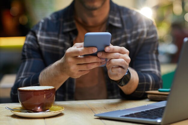 Close-up of man sitting at the table and typing a message on his mobile phone while drinking coffee at cafe