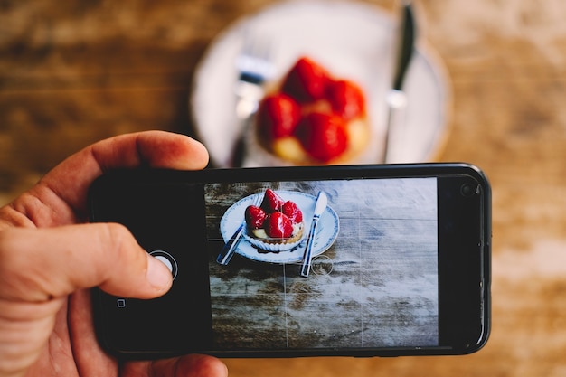 Close up of man taking picture of a little strawberry cake to share on social media and receive likes and followers. People and internet technology. Wooden table