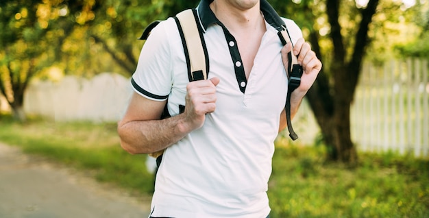 彼の肩にバックパックと白いTシャツで公園を歩いているクローズアップの男。暖かいカラー写真、白いフェンス、夏の天気