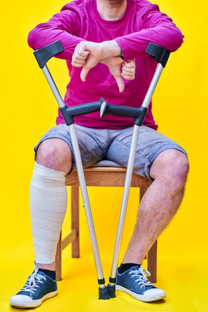 Photo close-up of a man with crutches, jeans and purple t-shirt sitting on a chair with his thumb down.