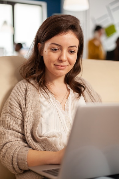 Close up of manager woman searching on laptop sitting in relax zone on confortable couch typing on pc smiling while diverse colleagues working in background