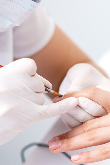 Close up of manicure master in rubber gloves applying transparent nail polish on female nails in beauty salon