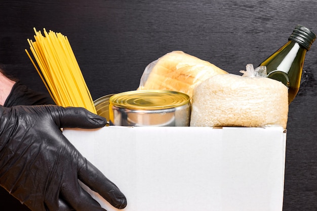 Close-up mans handen in zwarte rubberen handschoenen met een witte doos met pasta, granen en ingeblikt voedsel op een zwarte muur. veiligheid eten bezorgservice.