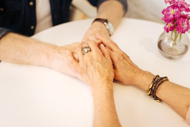 Close up of mature female and male hands holding each other and locating on white surface