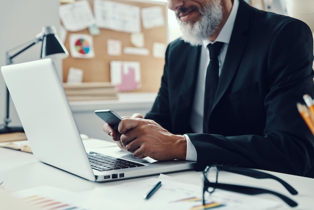 Close up of mature man in full suit smiling and using smart phone while working in modern office