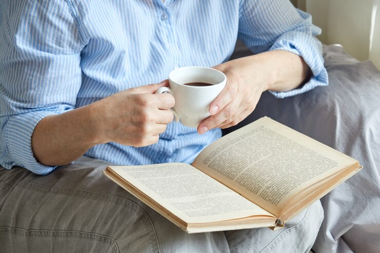コーヒーを飲みながらソファに座って、窓際の自宅で本を読んでいる成熟した女性のクローズアップ。