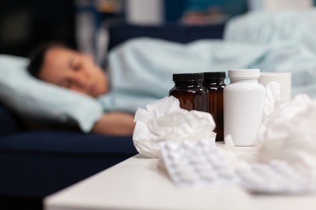 Close up of medication pills and napkins for sick woman sleeping on sofa taking drugs and medical tr...