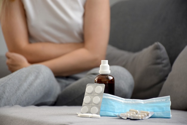 Close up on medicine, pills, tablets, syrup, protective mask and young woman holding her belly with hands and suffering from female pain