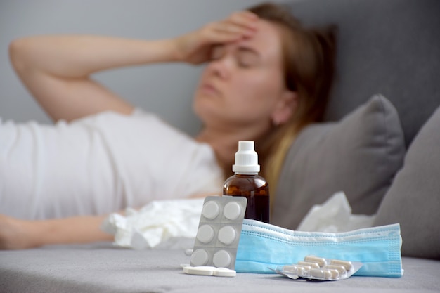 Close up on medicine, tablets, syrup, pills and protective mask and young woman touching her forehead with hand and suffering from headache