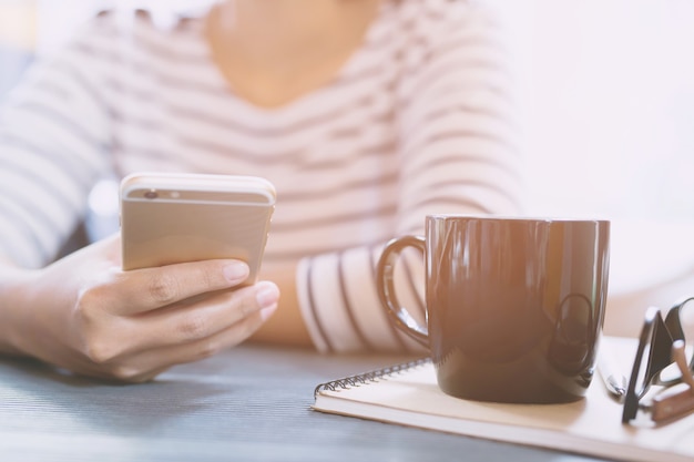 Close-up mensen vrouw zitten met behulp van mobiele smartphone in een pauze te ontspannen. kopje koffie op bureautafel met boek Kladblok, brillen, apparatuur benodigdheden in het werk. kopieer ruimte. concept werkende vrouw.