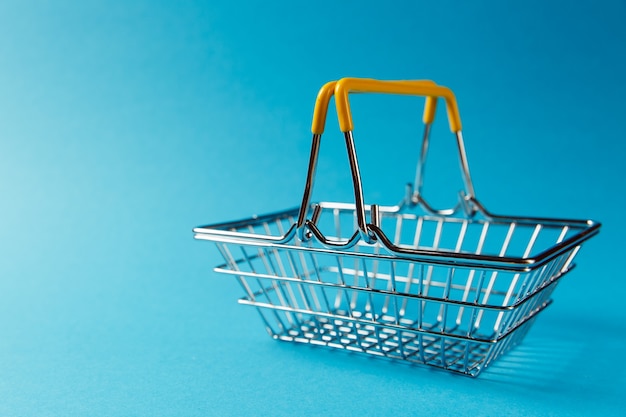 Close up of metal grocery basket for shopping in supermarket with raised handles and yellow plastic elements isolated on blue background. Concept of shopping. Copy space for advertisement.