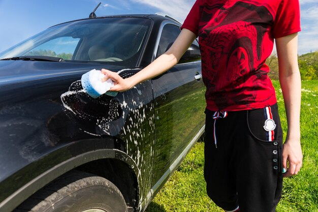 Close Up of Mid Section of Woman Wearing Athletic Clothing Washing Black Luxury Car with Soapy Sponge in Green Field on Bright Sunny Day