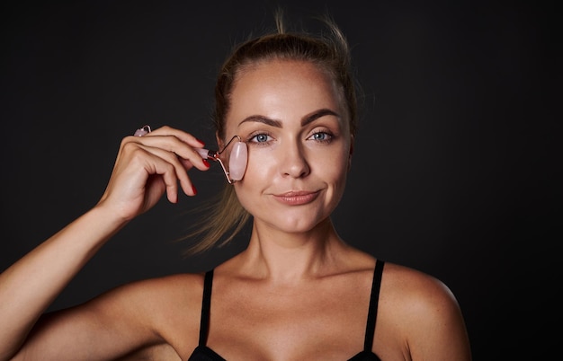 Close up of middle aged woman with healthy glowing tanned skin massaging face with jade roller isolated on black background Antiaging smoothing rejuvenation beauty therapy