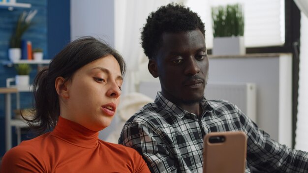 Primo piano di una coppia di razza mista che utilizza lo smartphone a casa nel soggiorno. persone sposate interrazziali che guardano il dispositivo sullo schermo e parlano mentre sono seduti sul divano. marito e moglie multietnici