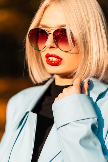 Close-up mode portret van mooie blonde vrouw met vintage zonnebril in trendy blauwe jas en trui op de natuur bij zonsondergang