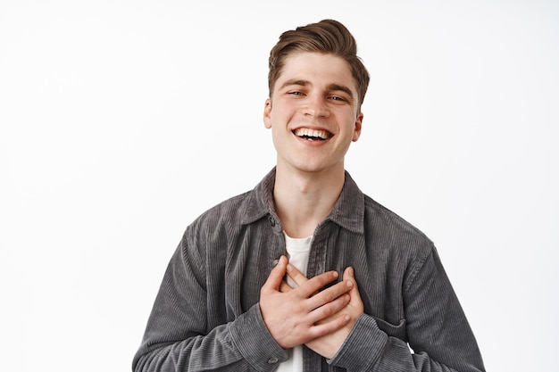 Close up of modern happy guy, young man laughing flattered, hold hands on chest heart and thank you, smiling pleased, delighted to recieve compliment, white background