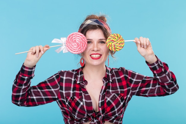 Foto close-up mooie lachende jonge vrouw in een geruit overhemd sluit haar ogen met grote kleurrijke lolly's en poseren tegen een blauwe achtergrond. concept van plezier en desserts