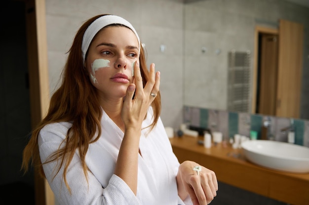 Close-up mooie roodharige Europese vrouw in witte badjas die hydraterend reinigend alginaatmasker toepast en zachtjes haar gezicht masseert en naar haar spiegelreflectie in de huisbadkamer kijkt