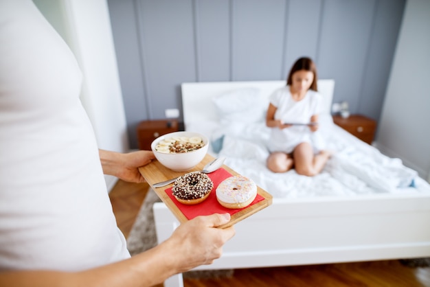 Close up of muscular man bringing doughnuts and fruit salad in the morning for his happy wife while sitting on the bed with a tablet in pajamas.