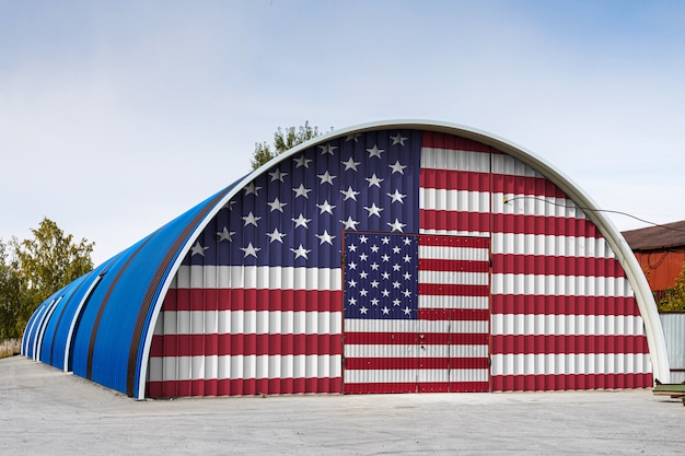 미국 국기의 근접 큰 창 고의 금속 벽에 푸른 하늘에 대하여 닫힌 된 영토를 그렸습니다.