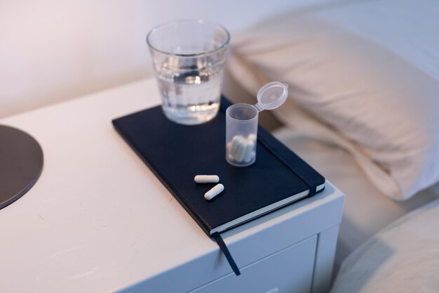 ベッドの隣のキャビネットに置かれたノート、錠剤の箱、水の入ったグラスの接写、新型コロナウイルスのパンデミックに対する隔離封鎖中の薬