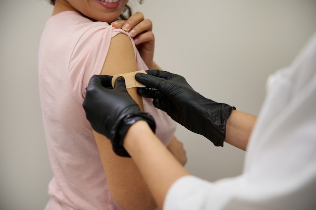 Primo piano delle mani di un'infermiera in guanti medici protettivi che mettono il cerotto, sulle mani di una donna sorridente felice di essere vaccinata per primi contro un'infezione virale da coronavirus.