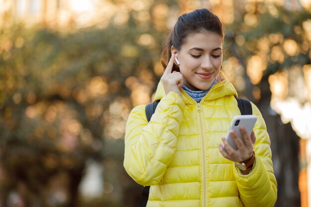 現代のワイヤレスイヤホンと街を歩いて彼女の携帯電話を使用して若いブルネットの女性のクローズアップ