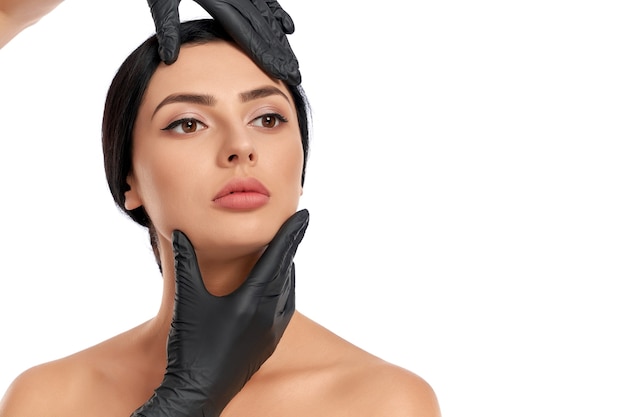 Крупным планом руки косметолога в черных медицинских перчатках, исследующих лицо женщины на белом