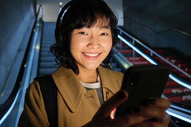 사진 헤드폰을 끼고 있는 귀여운 아시아 소녀가 에스컬레이터를 타고 내려가는 동안 들을 노래를 고른다