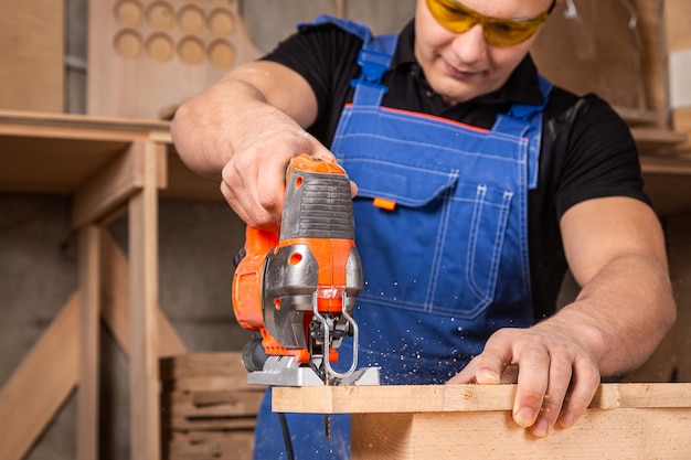 写真 熟練した木工が作業服を着て小規模なビジネスのオーナーである木工はワークショップで木製のバーのをジグソーで削り加工します