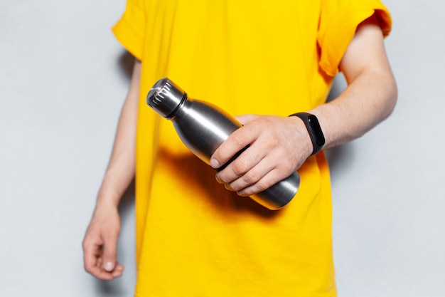 白い壁の背景に黄色のシャツを着て、鋼の再利用可能なサーモウォーターボトルを保持している男性の手のクローズアップ。