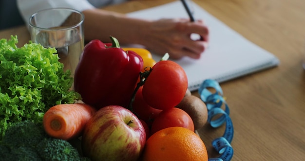 写真 果物や野菜でいっぱいのテーブルにダイエット計画を書いている栄養士の女性のクローズアップ健康的な栄養エコ食品