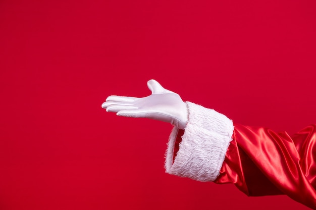 写真 赤い背景にジェスチャーを与えるサンタクロースの手袋をはめたハンドショーのクローズアップ。明けましておめでとう、メリークリスマス、伝統的な季節のお祝いのためのお祝いの時間。