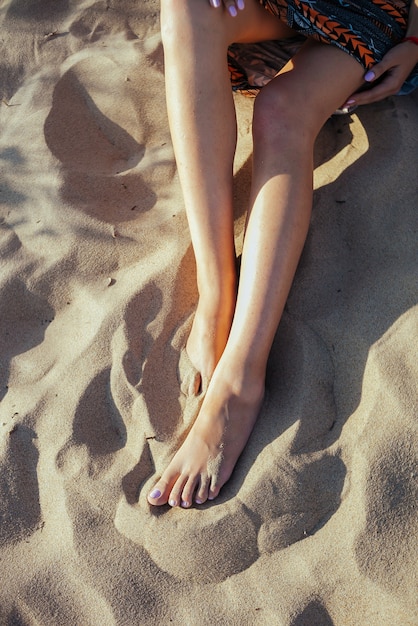 砂浜に裸足で座っている女性の足のクローズアップ。休暇、旅行、自由の概念。夏にリラックスする人々。