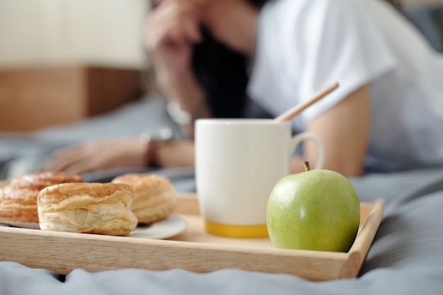 사진 노트북으로 일하는 남자가 있는 침대에 사과, 커피 머그, 빵과 같은 아침 식사가 포함된 나무 쟁반 클로즈업
