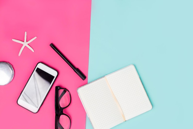 Close up office desk with stationery, glasses and smartphone. Spring pink and pastel blue background with womens work accessories