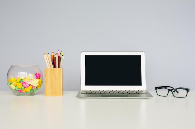 Close up of office desktop with blank laptop, spectacles, colorful pencils in wooden box and decorative glass bowl. Mock up