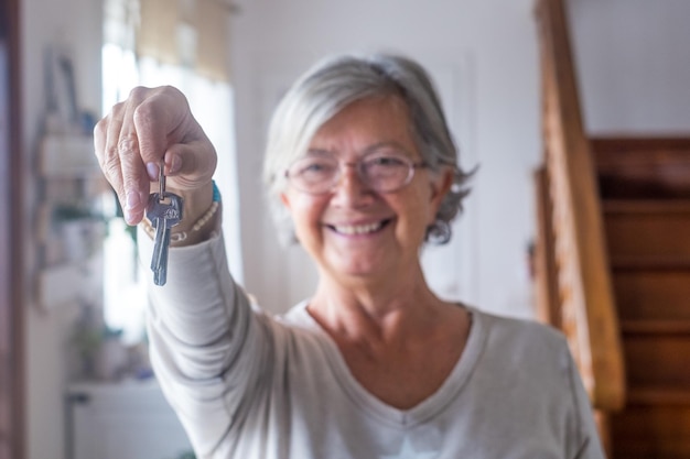 Close up of old and mature woman at home holding keys of house to the camera Portrait of female senior smiling and looking at the cameraxA