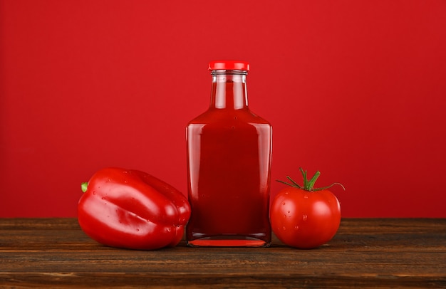 Photo close up one glass bottle of ketchup sauce, fresh tomatoes and bell pepper on wooden table over red background with copy space, low angle view