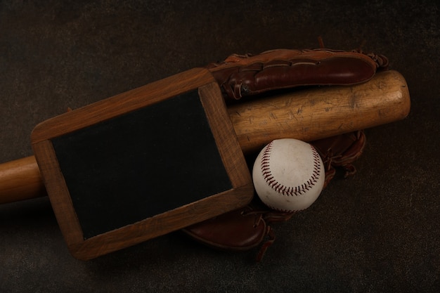 Close up one old baseball ball, wooden bat, worn leather vintage glove and blackboard sign on grunge dark brown background, elevated top view, directly above