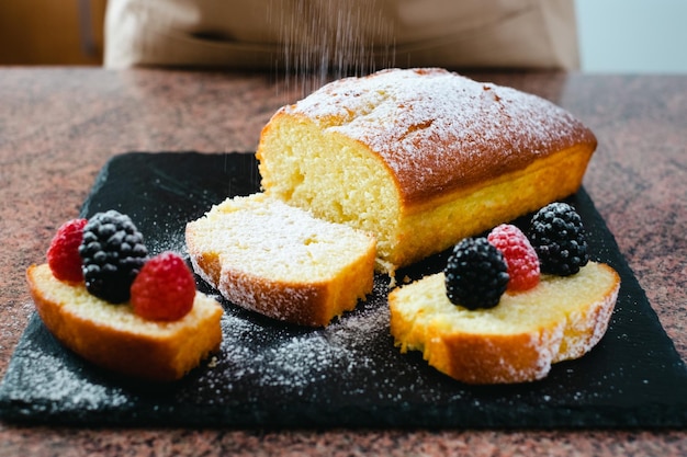 Close-up op de zelfgemaakte plumcake op een snijplank van zwarte steen terwijl hij wordt bestrooid met poedersuiker