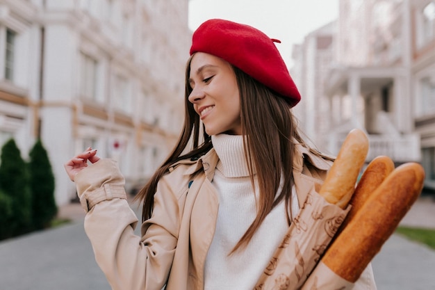 Close up outdoor photo of tender pretty girl in red beret and beige trench is smiling and looking aside with bagels